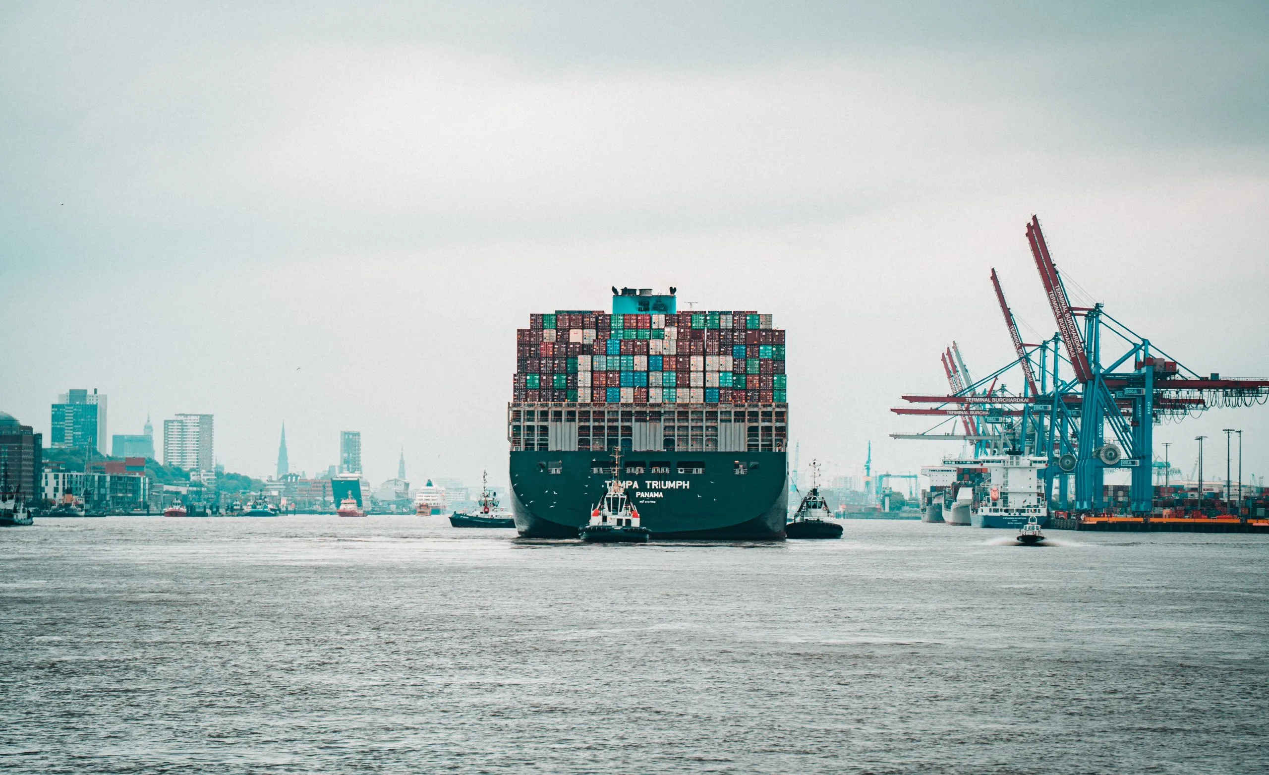 Container-Schiff im Hafen mit urbaner Skyline und Kränen im Hintergrund