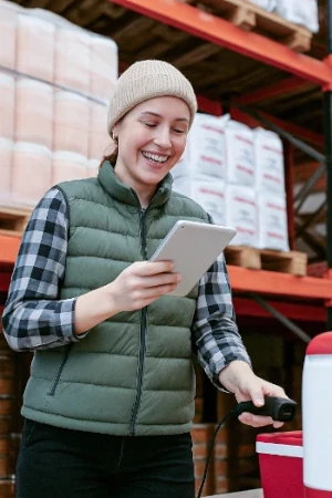 A smiling young woman in a beanie, plaid shirt, and vest uses a tablet and barcode scanner in a warehouse setting.